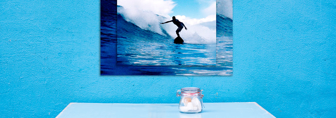 A canvas print of a surfer riding a wave, mounted on a bright blue textured wall above a small table with a jar