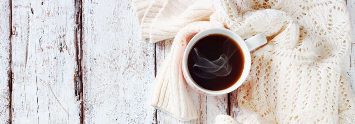A steaming mug of dark coffee resting on a cream knit scarf atop a white rustic wooden table